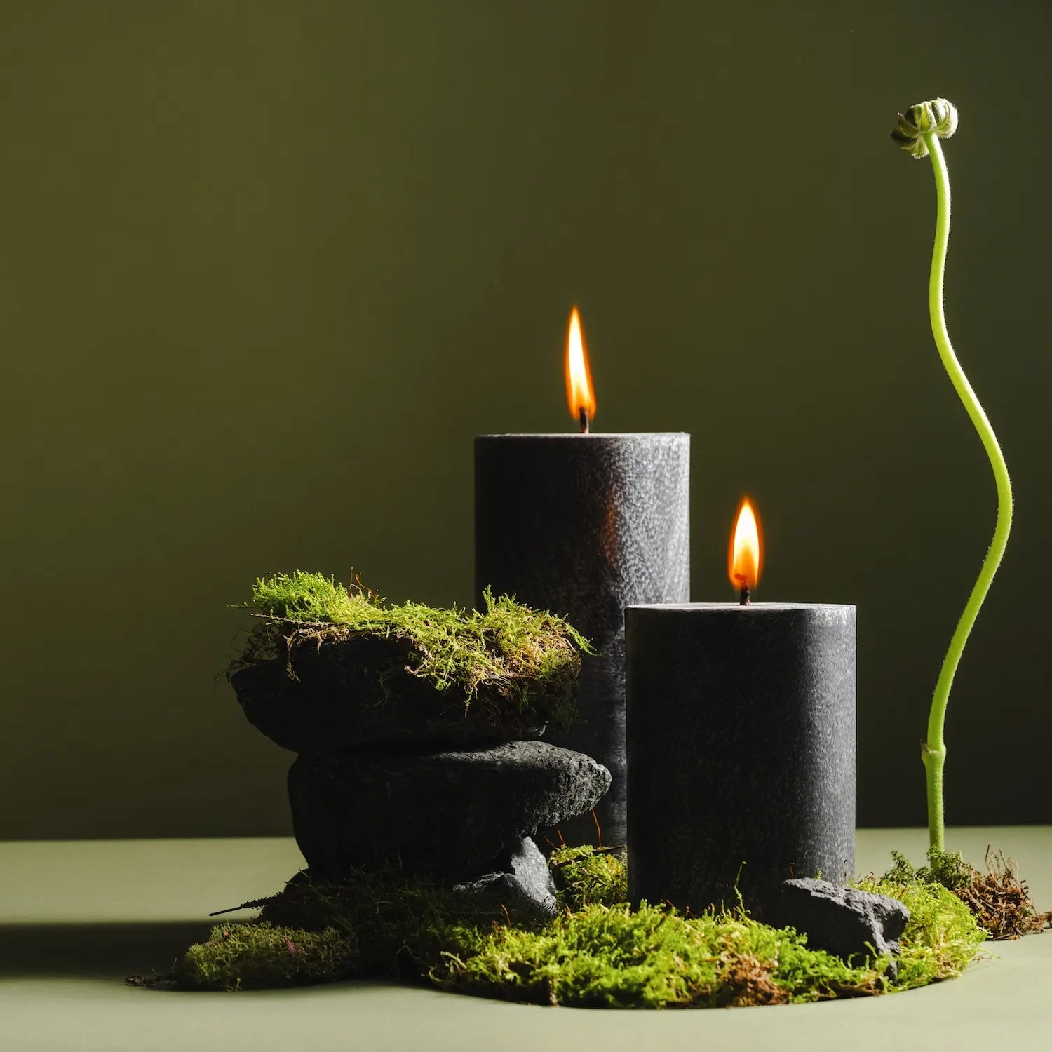Two lit black pillar candles with small orange flames sit on stacked dark stones and bright green moss, next to a tall, curved plant shoot, set against a soft olive-green background.