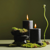 Two lit black pillar candles with small orange flames sit on stacked dark stones and bright green moss, next to a tall, curved plant shoot, set against a soft olive-green background.