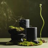 Two black cylindrical candles releasing wisps of smoke, arranged on dark stones covered with green moss, beside a slender green plant shoot curving upward against a muted olive background.