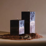 Three rectangular black candles with marbled gray-white sides stand unlit in a mound of reddish-brown soil, with a small stone in front, set against a warm neutral background.