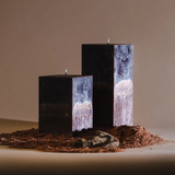 Three rectangular black candles with marbled gray-white sides stand unlit in a mound of reddish-brown soil, with a small stone in front, set against a warm neutral background.