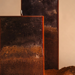 Close-up of two tall rectangular candles with layered dark brown, rust, and charcoal tones, showing a textured, stone-like surface, set against a soft beige background with earthy soil at the base.