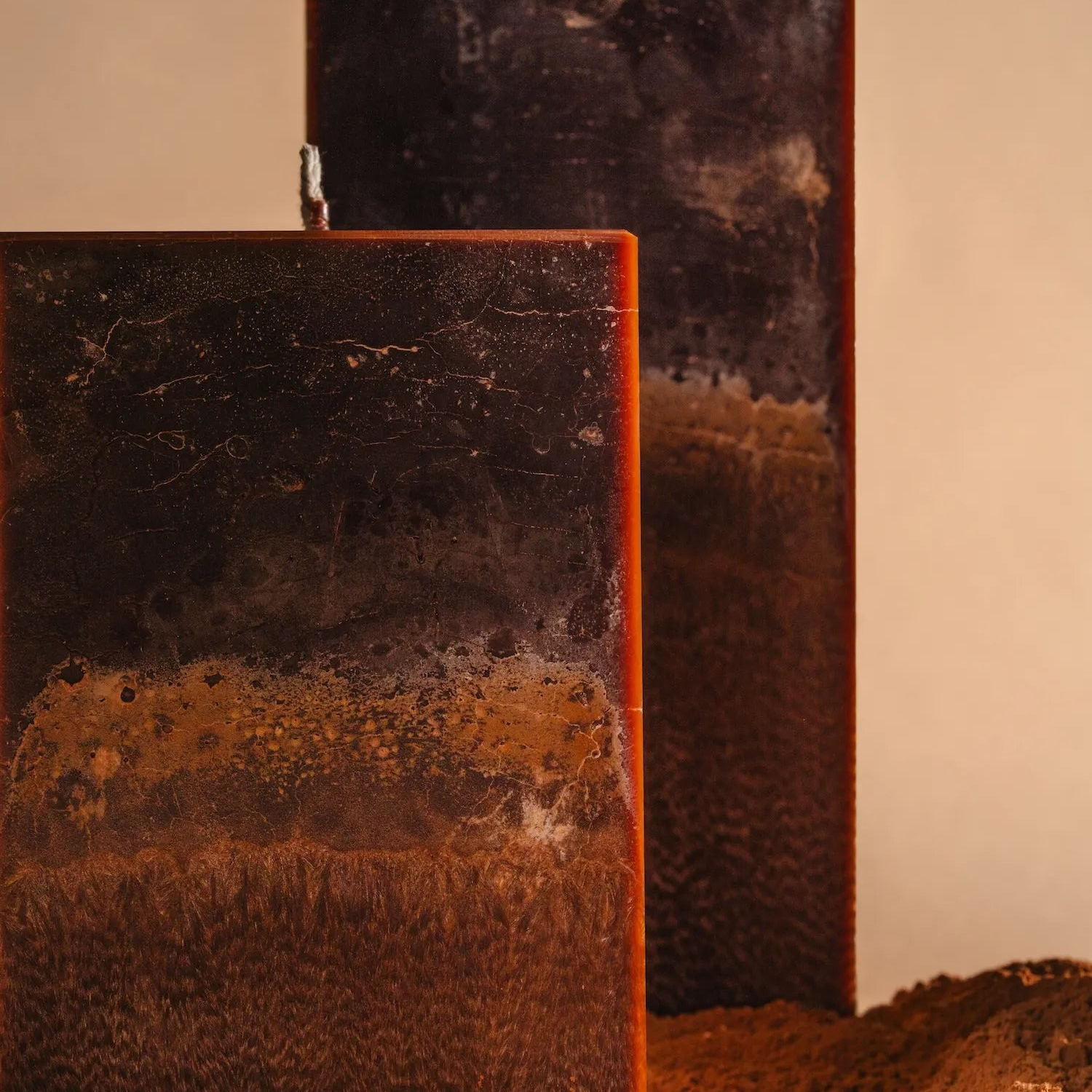 Close-up of two tall rectangular candles with layered dark brown, rust, and charcoal tones, showing a textured, stone-like surface, set against a soft beige background with earthy soil at the base.