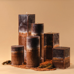 A sculptural arrangement of unlit candles in varied heights and shapes-rectangular and cylindrical-with layered dark brown, rust, and charcoal coloring, set in reddish soil with small stones against a warm beige background.