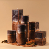 A sculptural arrangement of unlit candles in varied heights and shapes-rectangular and cylindrical-with layered dark brown, rust, and charcoal coloring, set in reddish soil with small stones against a warm beige background.