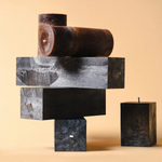 An abstract stack of unlit candles in rectangular and cylindrical forms, layered in dark charcoal, gray, and rust tones, balanced like blocks against a soft beige background.